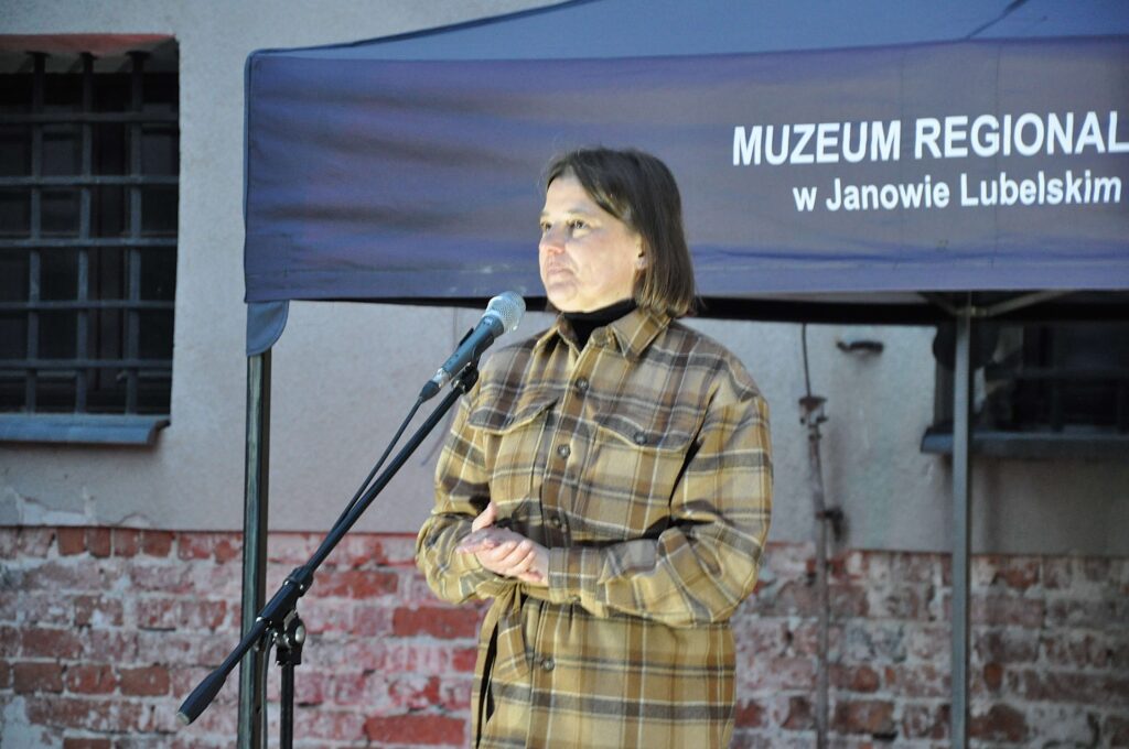 Kobieta w brązowej, kraciastej kurtce stoi przy mikrofonie na tle ściany z cegły i namiotu z napisem Muzeum Regionalne.