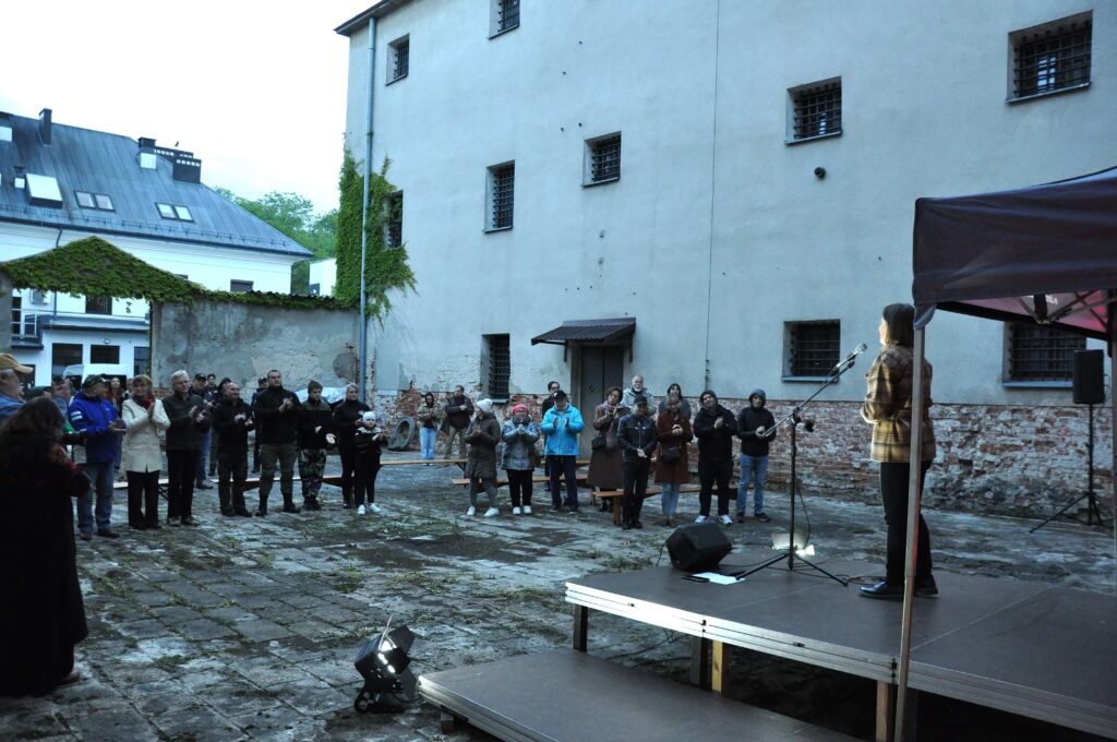 Grupa ludzi stojących na kamiennym dziedzińcu przed podwyższoną sceną, na której stoi kobieta przy mikrofonie, szare mury budynków w tle.