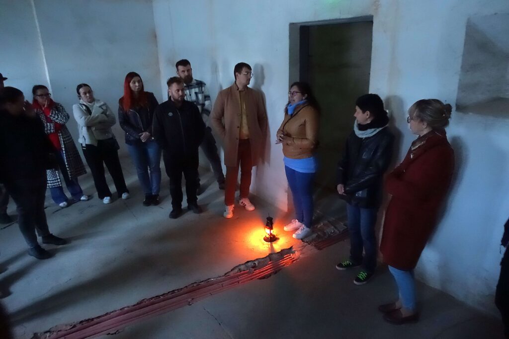 Grupa ludzi stoi w półokręgu w ciemnym, surowym pomieszczeniu wokół zapalonej lampy na podłodze.