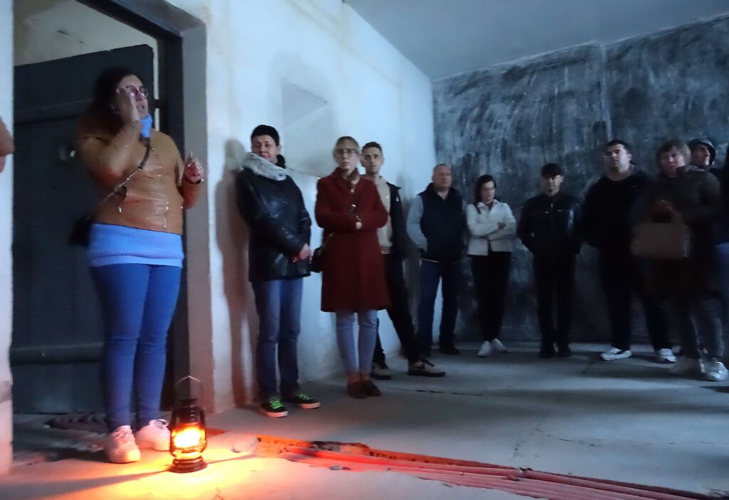 Grupa osób stojąca w ciemnym pomieszczeniu przy ścianie, na podłodze świeci się lampka naftowa.