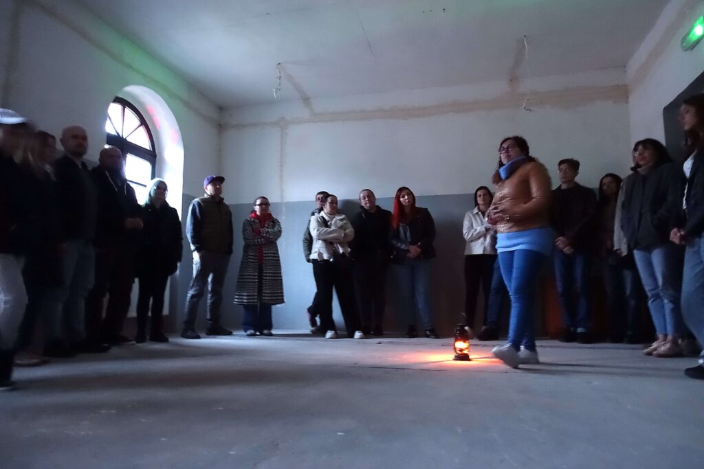 Grupa ludzi stoi w półokręgu w dużym pustym pomieszczeniu z jasnoszarymi ścianami, na środku pomieszczenia świeci mała pomarańczowa lampa.