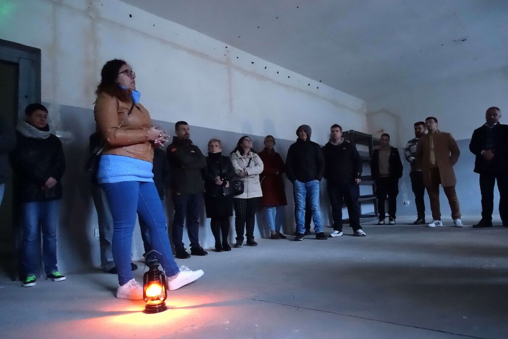 Grupa ludzi w ciemnym pomieszczeniu, jedna kobieta z brązową kurtką i niebieskimi spodniami stoi obok świecącej lampy na podłodze.