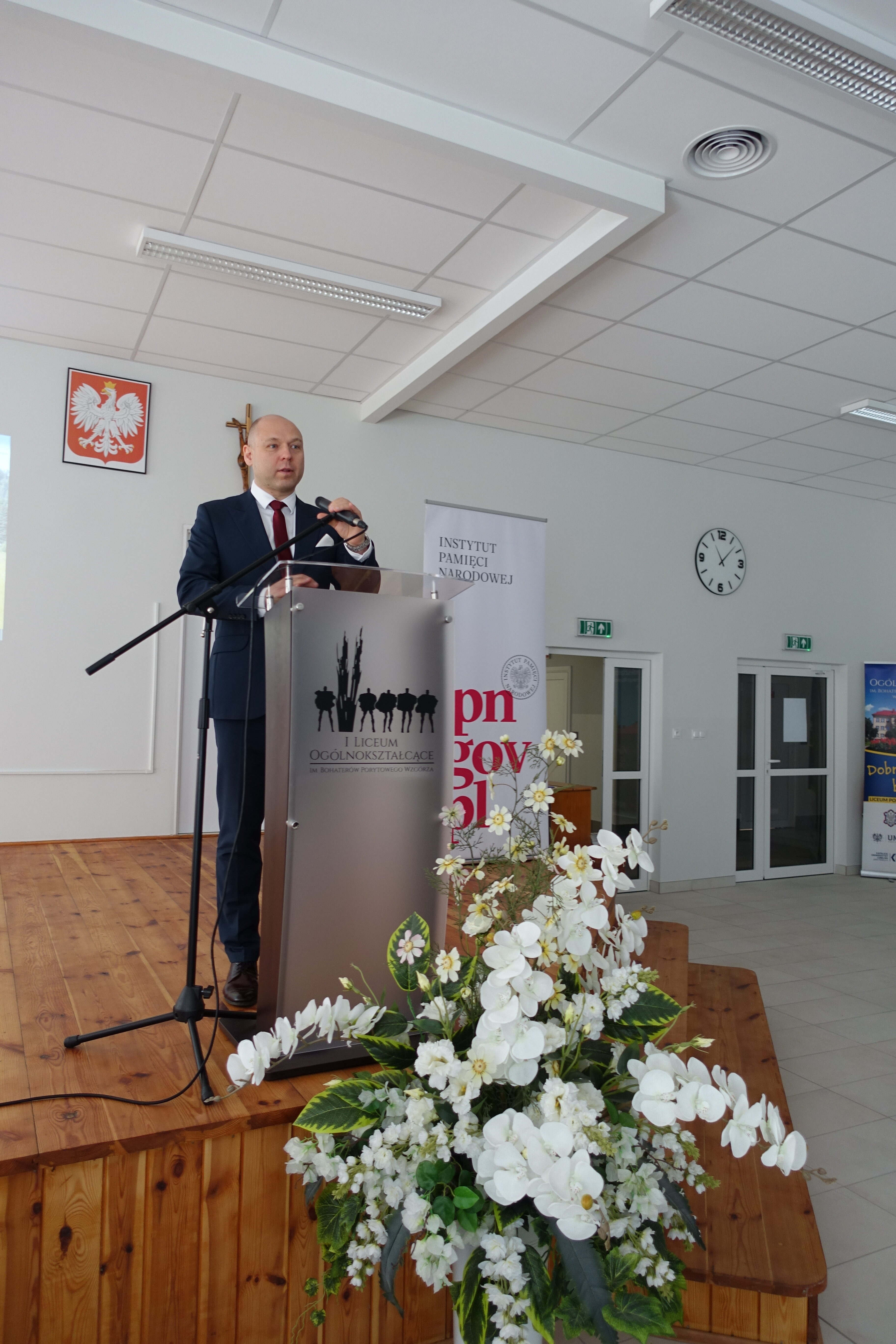 Mężczyzna w granatowym garniturze przemawia na drewnianej scenie za szarą mównicą z logo szkoły, obok bukiet białych kwiatów.