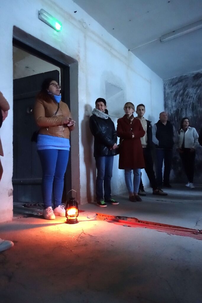 Grupa osób stojąca przy ścianie w ciemnym pomieszczeniu, na podłodze stoi zapalona lampa naftowa emitująca pomarańczowe światło.