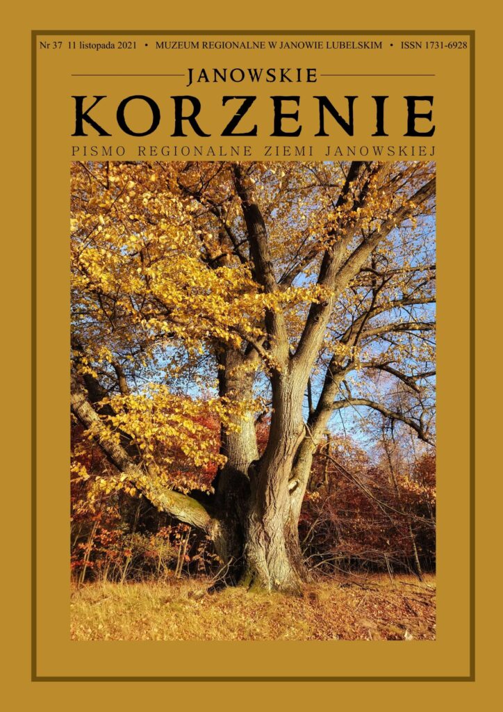 Stare drzewo z grubym pniem i gałęziami, żółte i pomarańczowe liście, trawa i las w jesiennych kolorach pod niebieskim niebem.