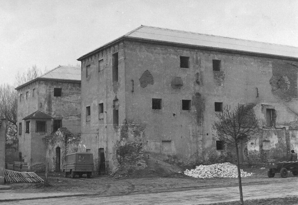 Stare, zniszczone budynki z cegły i tynku, sterta gruzu, bezlistne drzewo i dwa pojazdy na ziemnym podłożu.