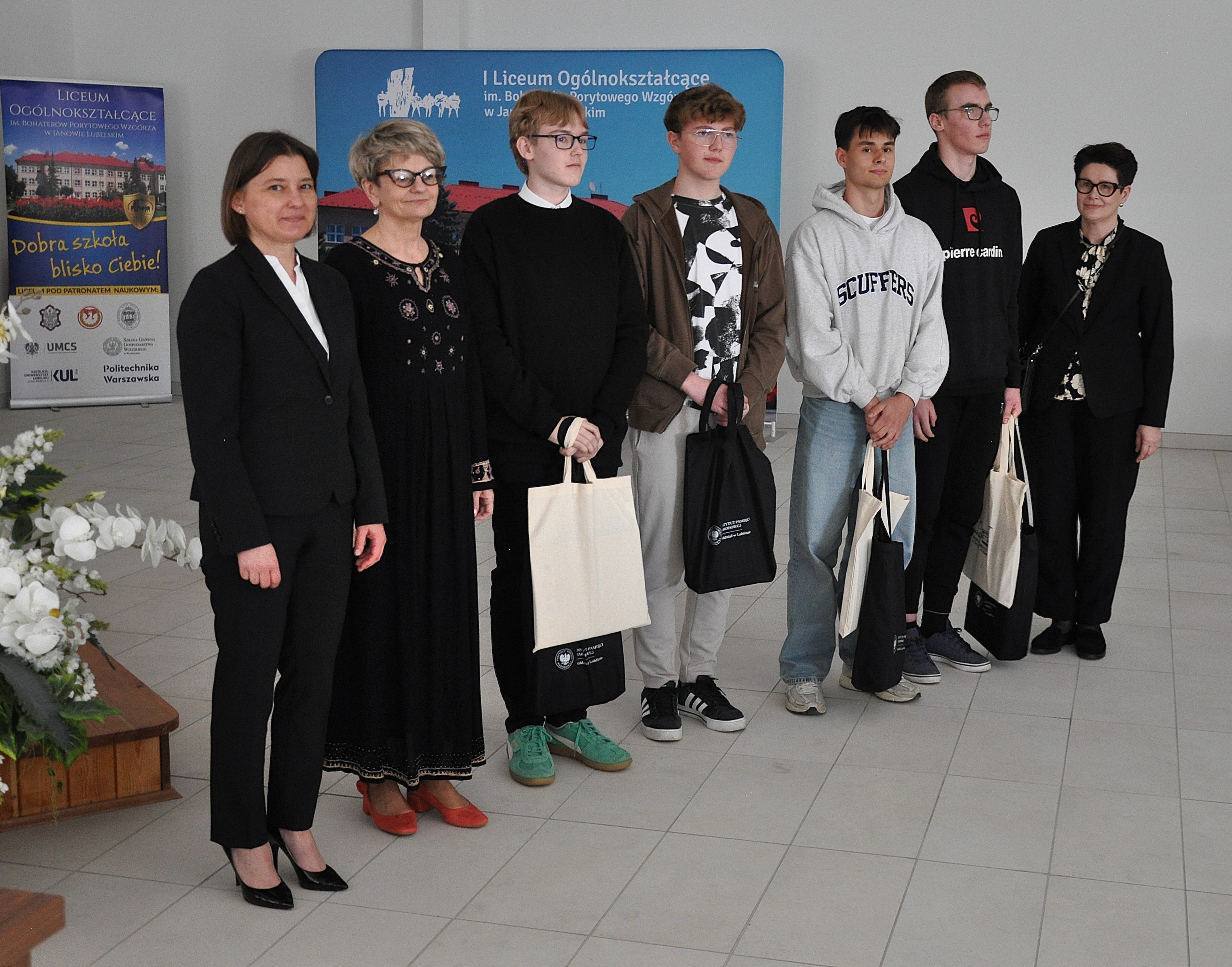 Siedem osób stoi w rzędzie na jasnej podłodze, w tle banery z napisem I Liceum Ogólnokształcące, większość trzyma torby bawełniane.