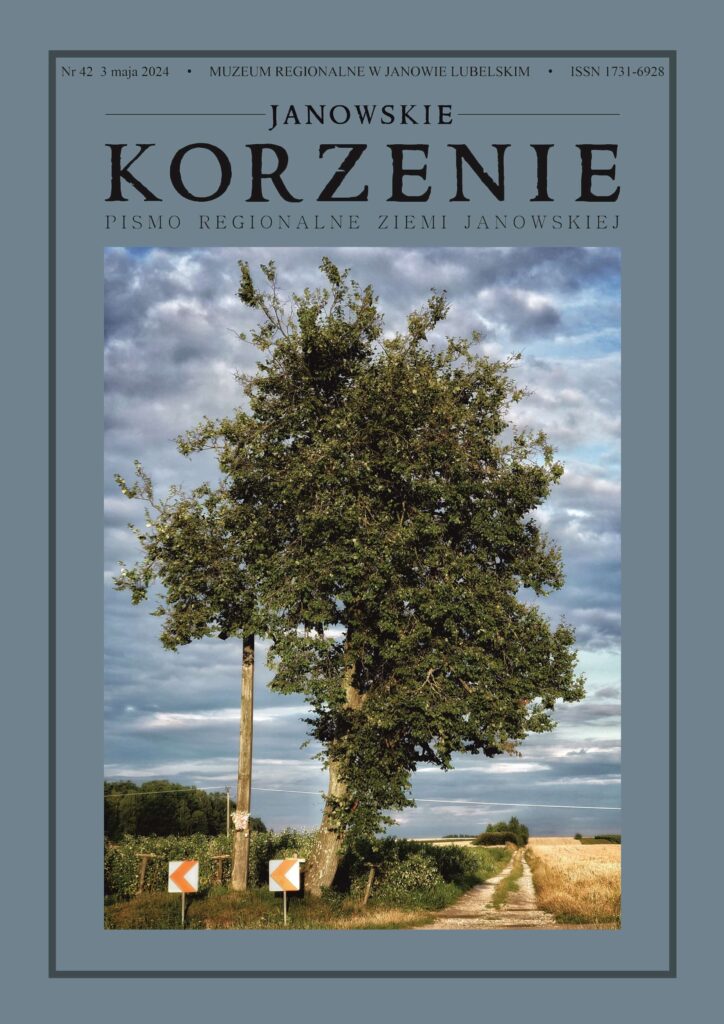 Drzewo przy wiejskiej drodze, obok słup z kwiatami i znaki kierunkowe, pola i chmurzyste niebo.