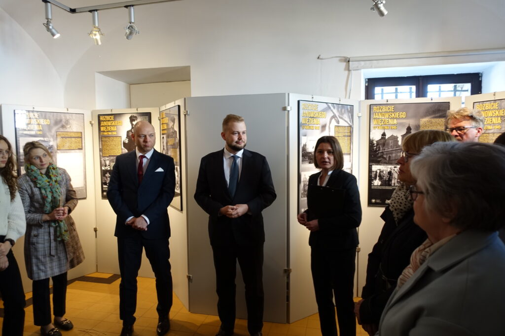 Grupa ludzi w formalnych ubraniach stoi w galerii z planszami wystawy o rozbiciu janowskiego więzienia.