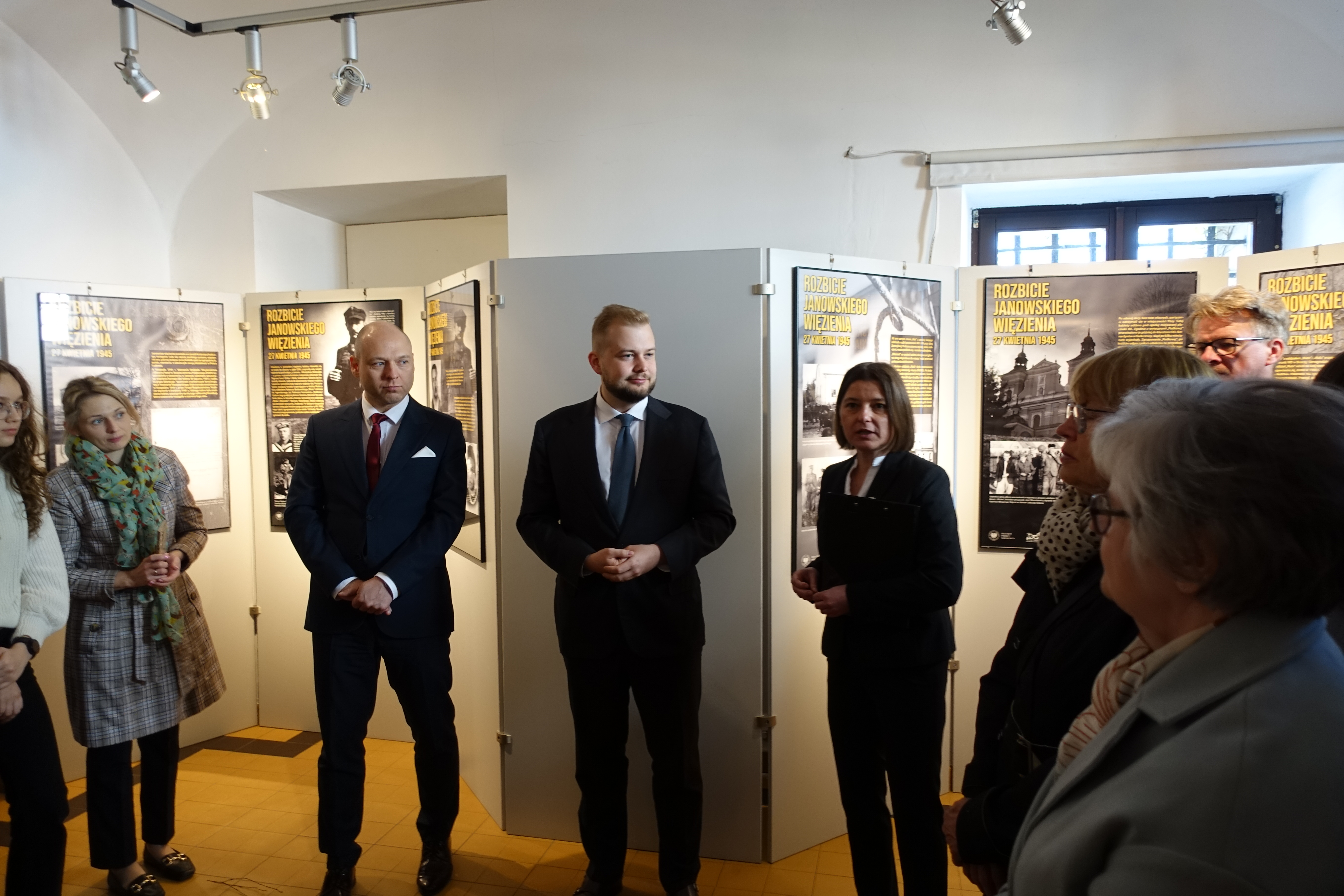 Grupa ludzi w formalnych ubraniach stoi w galerii z planszami wystawy o rozbiciu janowskiego więzienia.