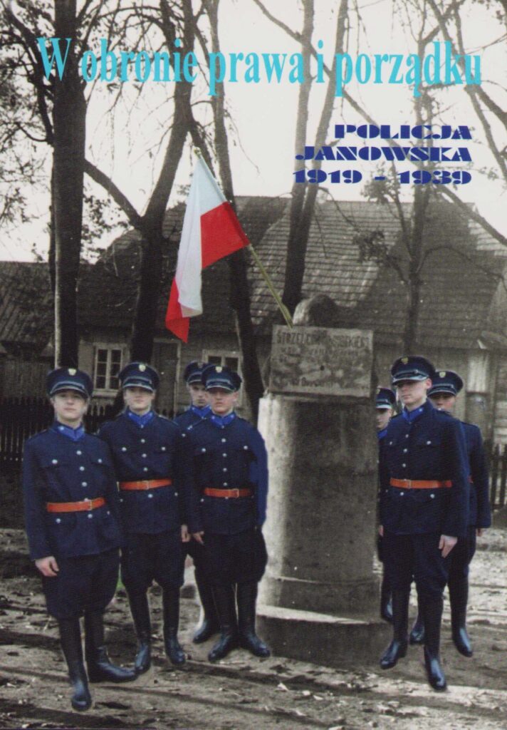 Mężczyźni w granatowych mundurach z czerwonymi pasami stojący obok kamiennego słupa z biało-czerwoną flagą, drzewa i budynki w tle.