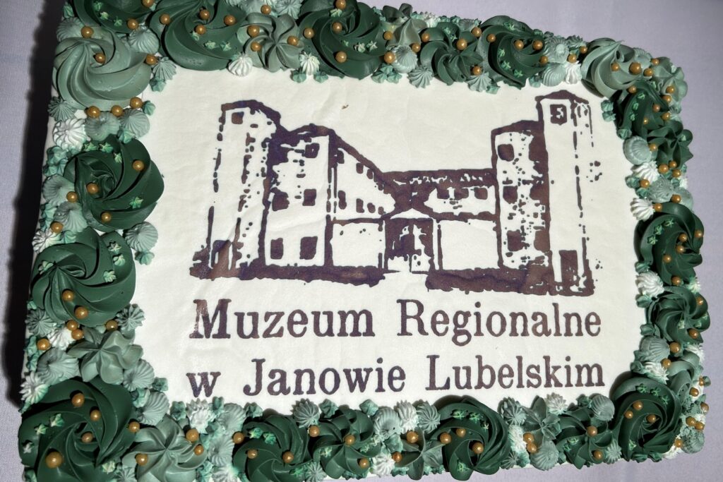 Prostokątny tort z białym kremem, zielone i białe dekoracje kremowe, złote kulki, nadruk budynku i napis "Muzeum Regionalne w Janowie Lubelskim".