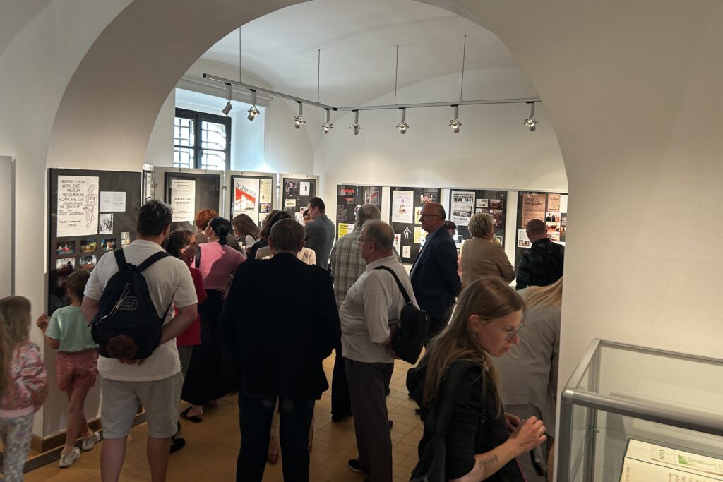 Grupa ludzi ogląda wystawę muzealną w jasnym pomieszczeniu z łukowym wejściem i tablicami z dokumentami na ścianach.