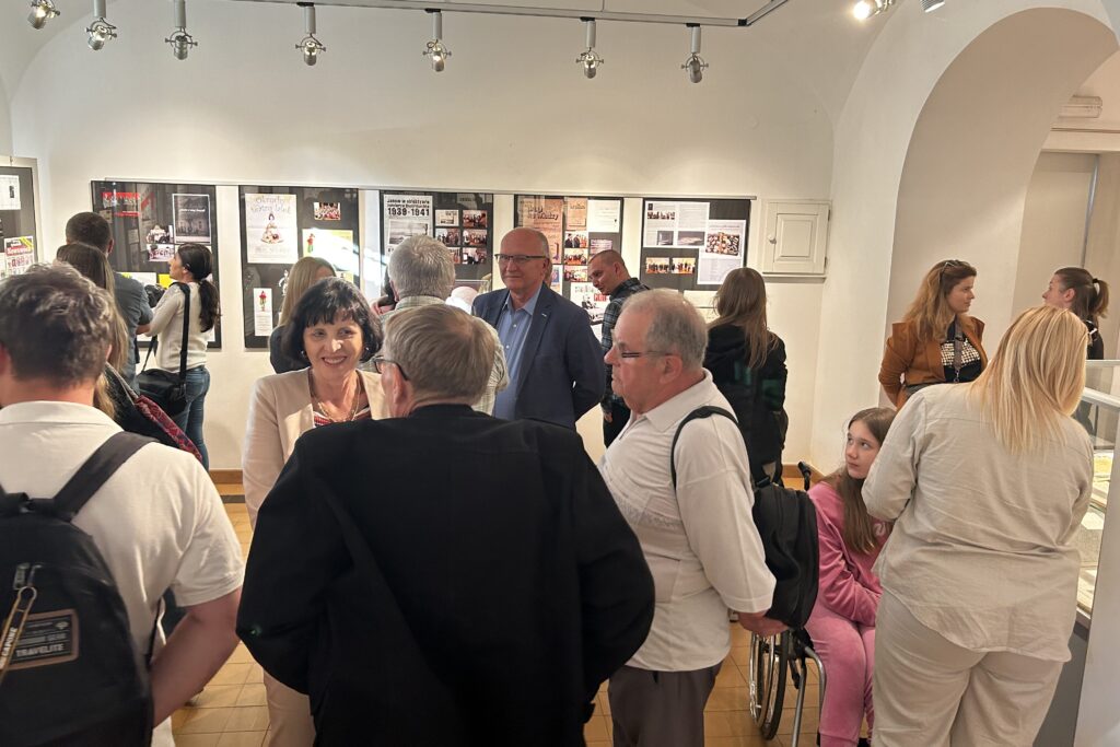 Grupa ludzi w jasnym pomieszczeniu z białymi ścianami, na których wiszą plansze z tekstami i zdjęciami, część osób rozmawia, dziewczynka na wózku w różowym ubraniu.