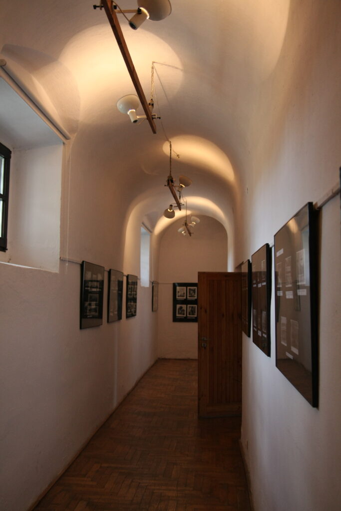 Wąski korytarz z białymi ścianami, drewnianą podłogą, drewnianymi drzwiami i czarnymi ramami obrazów oraz lampami sufitowymi.